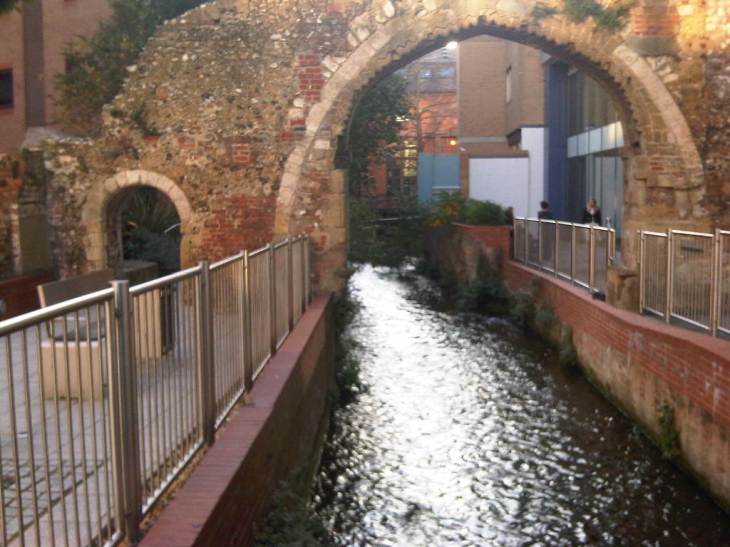 A stream runs through a narrow culvert between modern brick walls, with shiny metal rails, but flows beneath an ancient archway.  Behind the archway large, modern buildings close in.